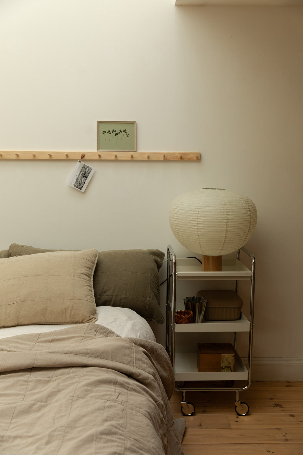 Calm Scandinavian bedroom with paper globe table lamp on chrome trolley, linen bedding and botanical print on peg rail