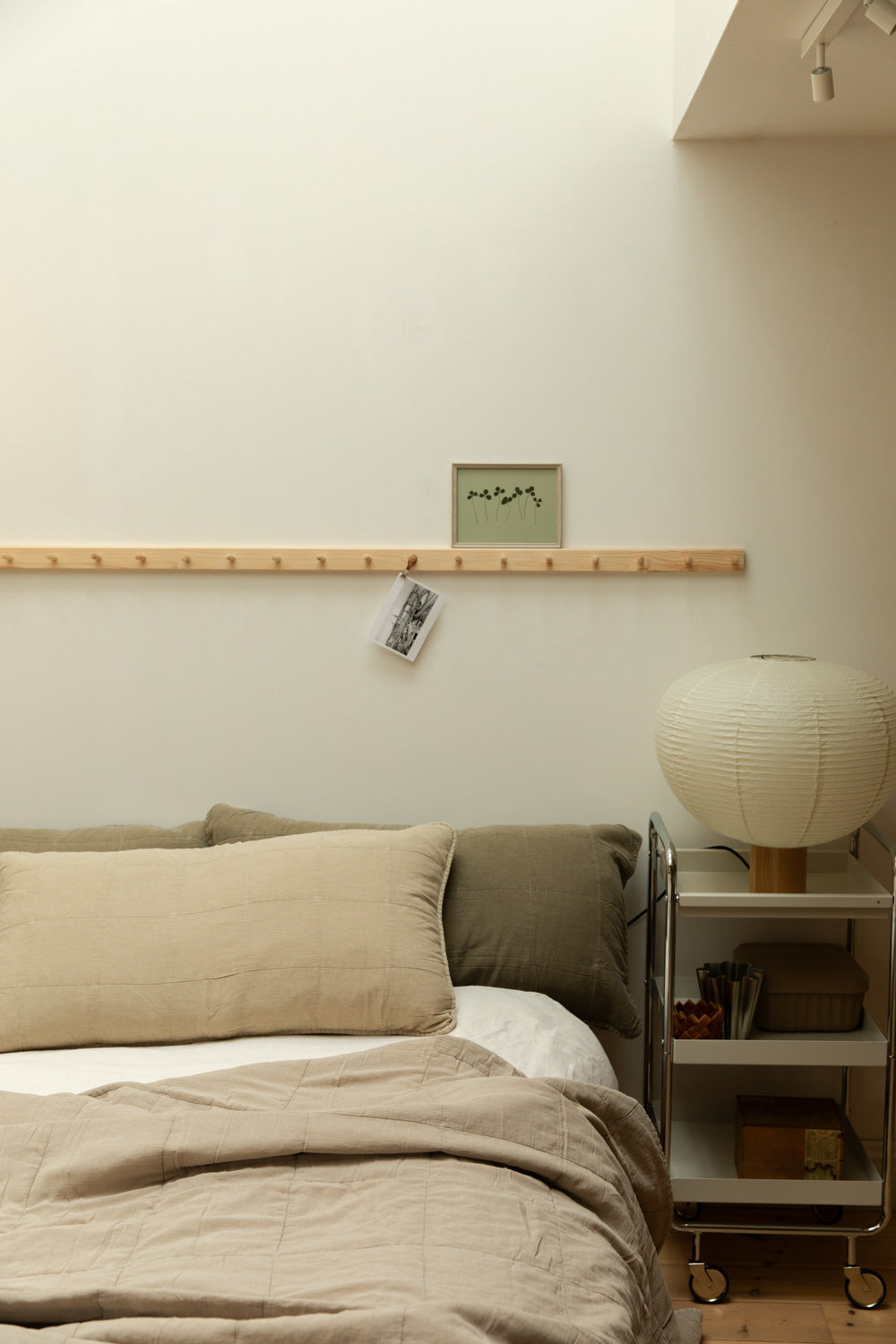 Calm Scandinavian bedroom with paper globe table lamp on chrome trolley, linen bedding, fog headboard cushion resting against two eucalyptus headboard cushions and botanical print on peg rail.