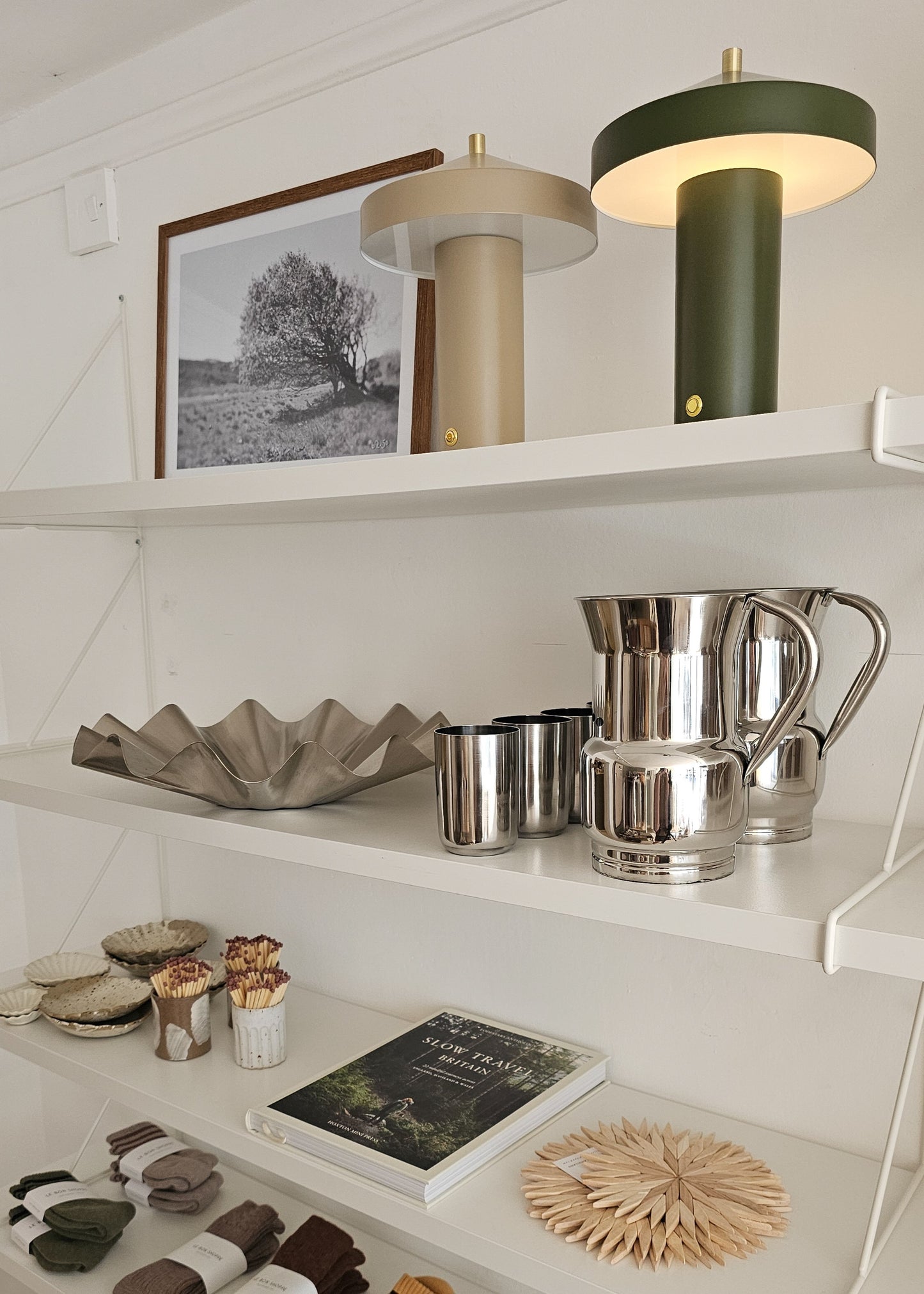 A styled interior shelf display featuring two modern table lamps, a framed black-and-white landscape print, a metallic bowl, silver pitchers and cups, small ceramic vessels, a book, and a woven coaster against a white wall.