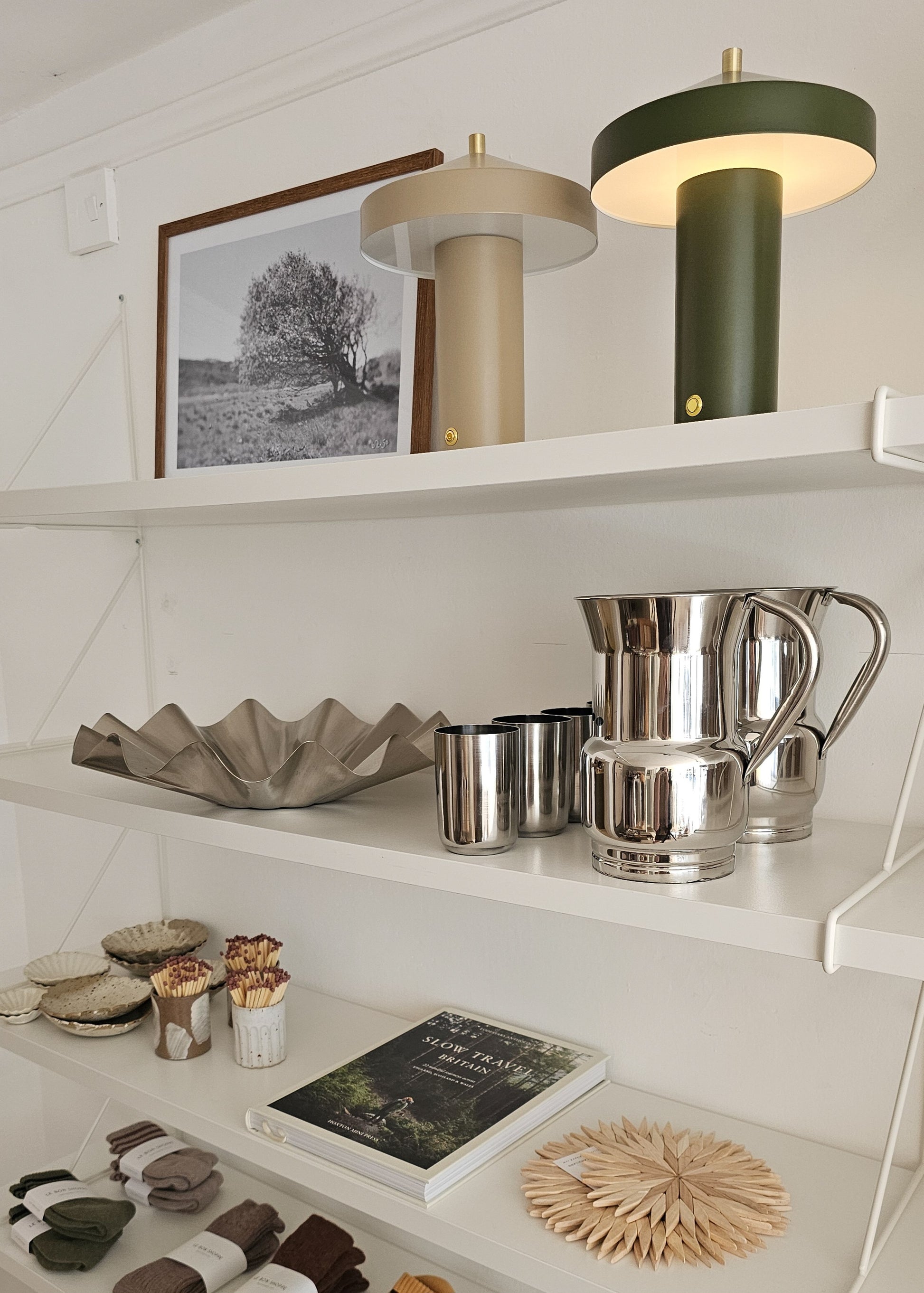 A styled interior shelf display featuring two modern table lamps, a framed black-and-white landscape print, a metallic bowl, silver pitchers and cups, small ceramic vessels, a book, and a woven coaster against a white wall.