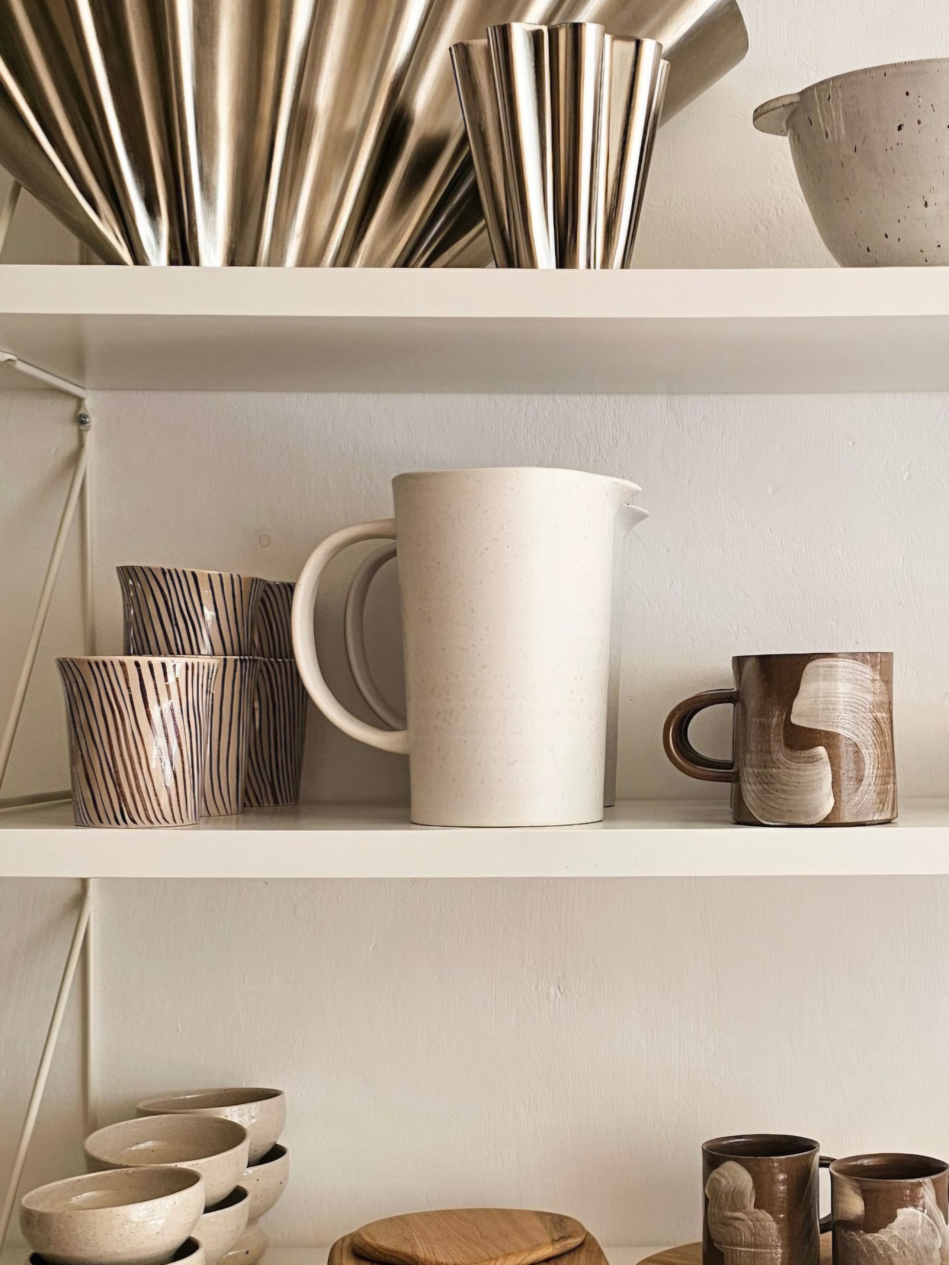 A styled interior shelf display featuring ceramic and metallic homeware, including a large off-white pitcher, patterned cups, bowls, and brushed metal vessels against a warm neutral wall.