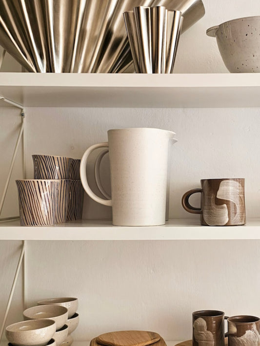 A styled interior shelf display featuring ceramic and metallic homeware, including a large off-white pitcher, patterned cups, bowls, and brushed metal vessels against a warm neutral wall.