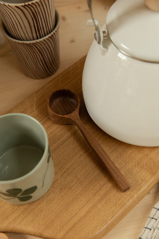 Selwyn House handmade English walnut coffee scoop on an oak board beside a white enamel kettle, Diva stripe mugs and a green floral mug.