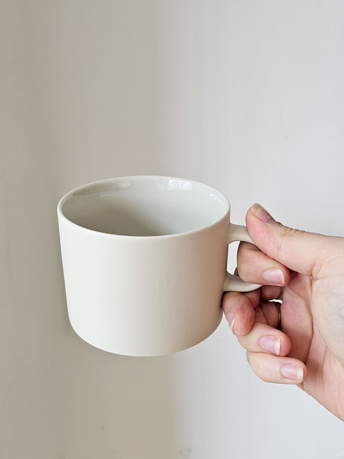 A hand holding a minimalist ceramic mug with a wide, rounded body and a small loop handle, shown against a plain light beige background.
