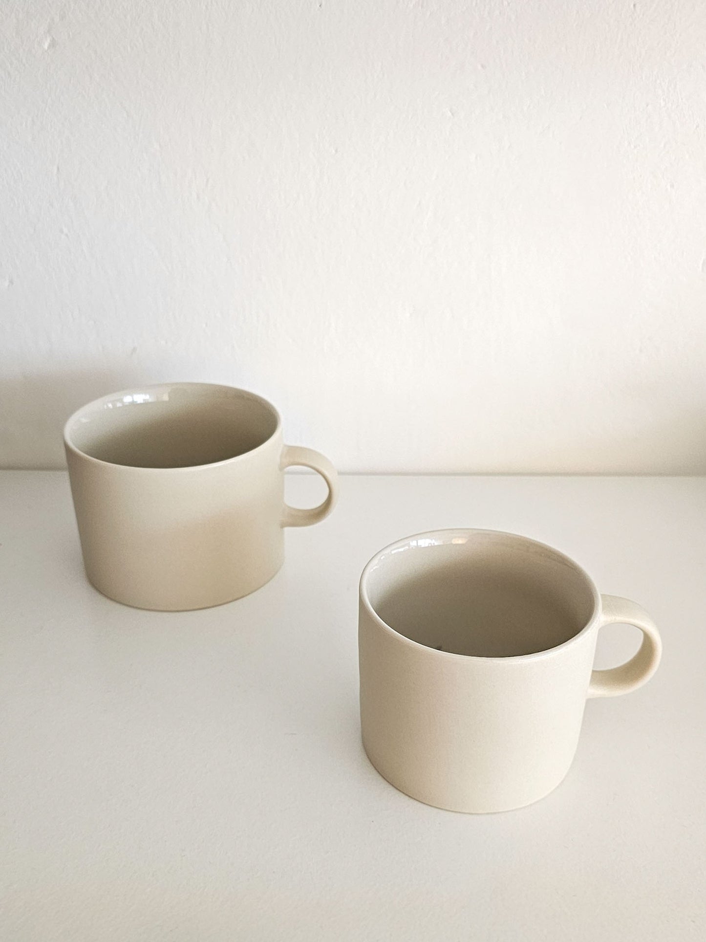 Two minimalist ceramic mugs in a warm off-white or cream color, displayed on a white surface against a white wall.