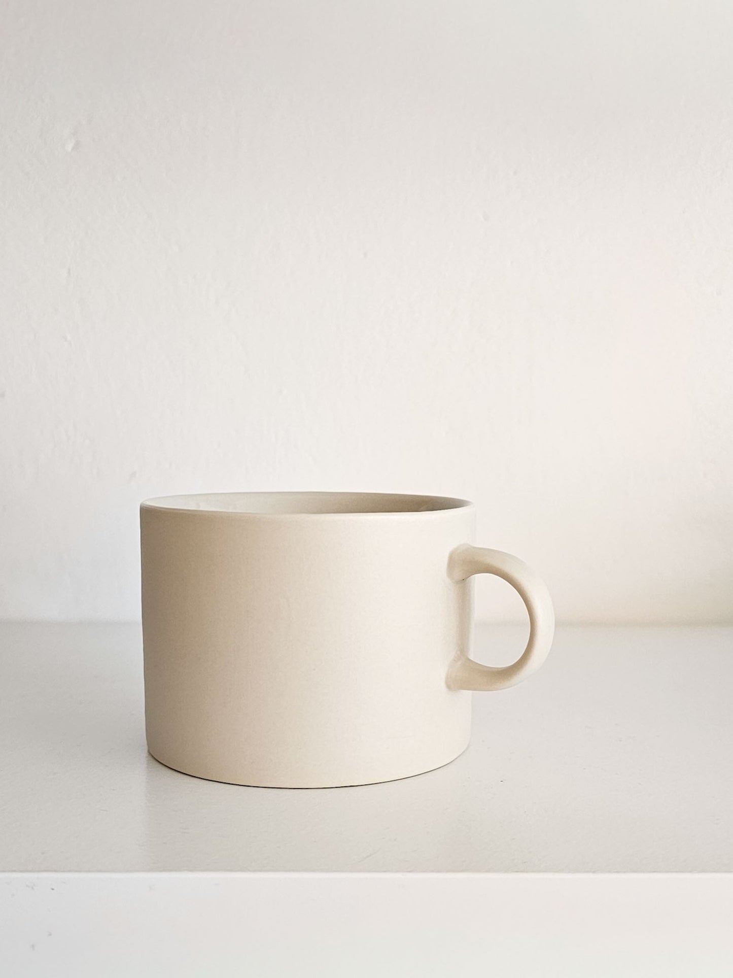 A minimalist ceramic mug in a warm cream or off-white color, featuring a simple loop handle and a matte finish, set against a clean, neutral white background.