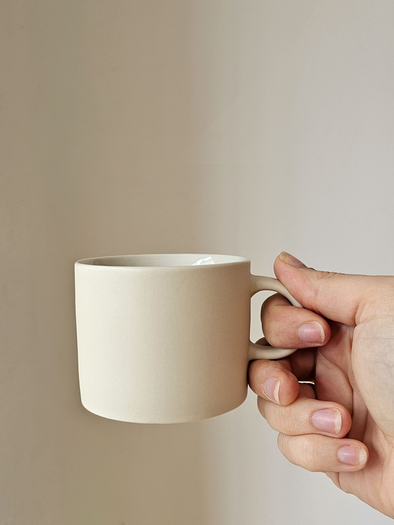 A hand holding a matte ceramic mug in a warm off-white or cream color against a plain beige background.