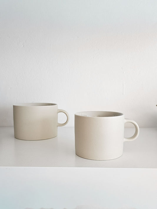 Two matte ceramic mugs in a warm off-white or cream color, sitting on a white shelf against a white wall.