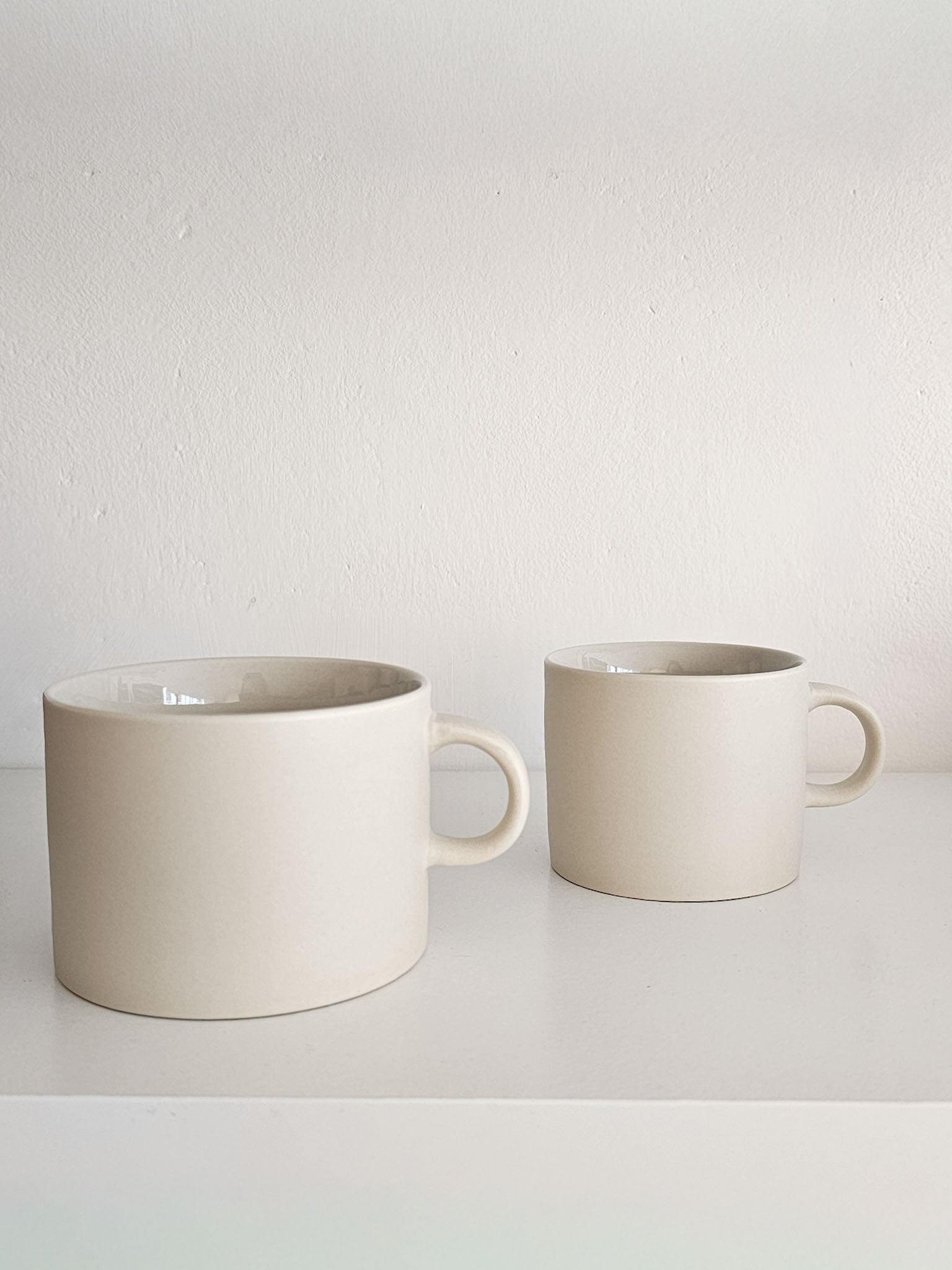 Two matte ceramic mugs in a warm off-white or cream color, displayed on a white shelf against a textured white wall.
