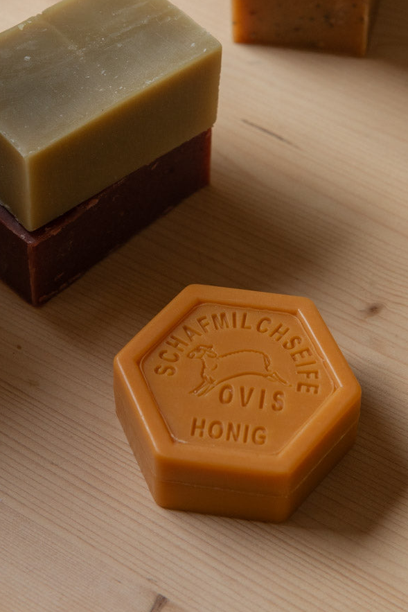 Hexagonal sheep's milk honey soap in front of stacked soap bricks and a wooden brush