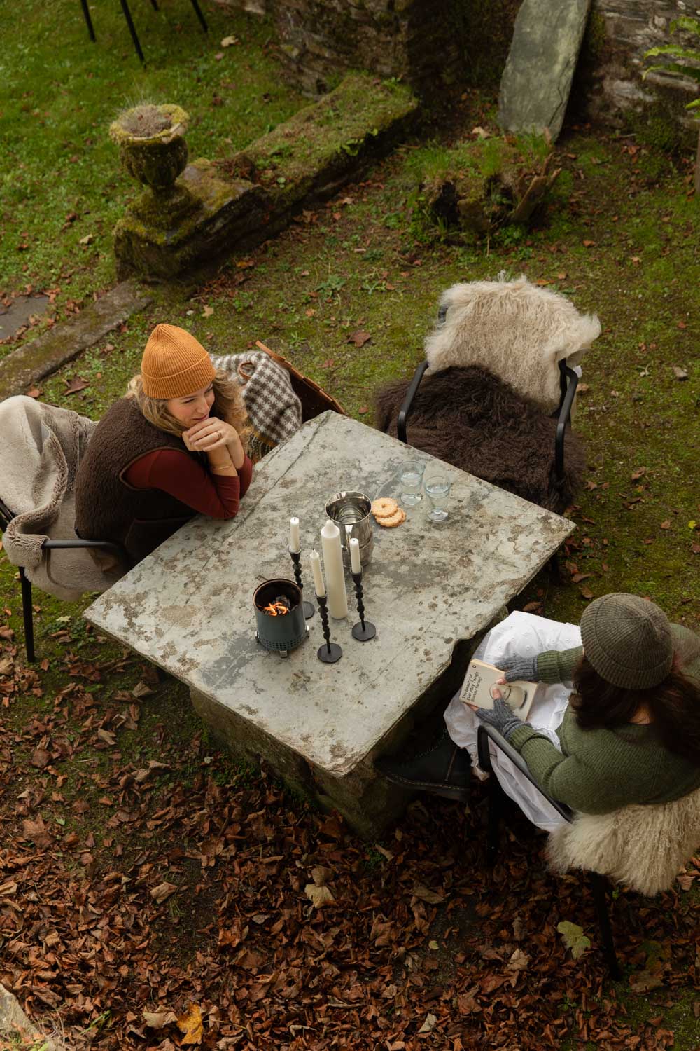 Overhead view of two women in Goldcrest and Granite Heather wool beanies at a stone garden table set with candles, wrapped in sheepskins and a houndstooth wool blanket