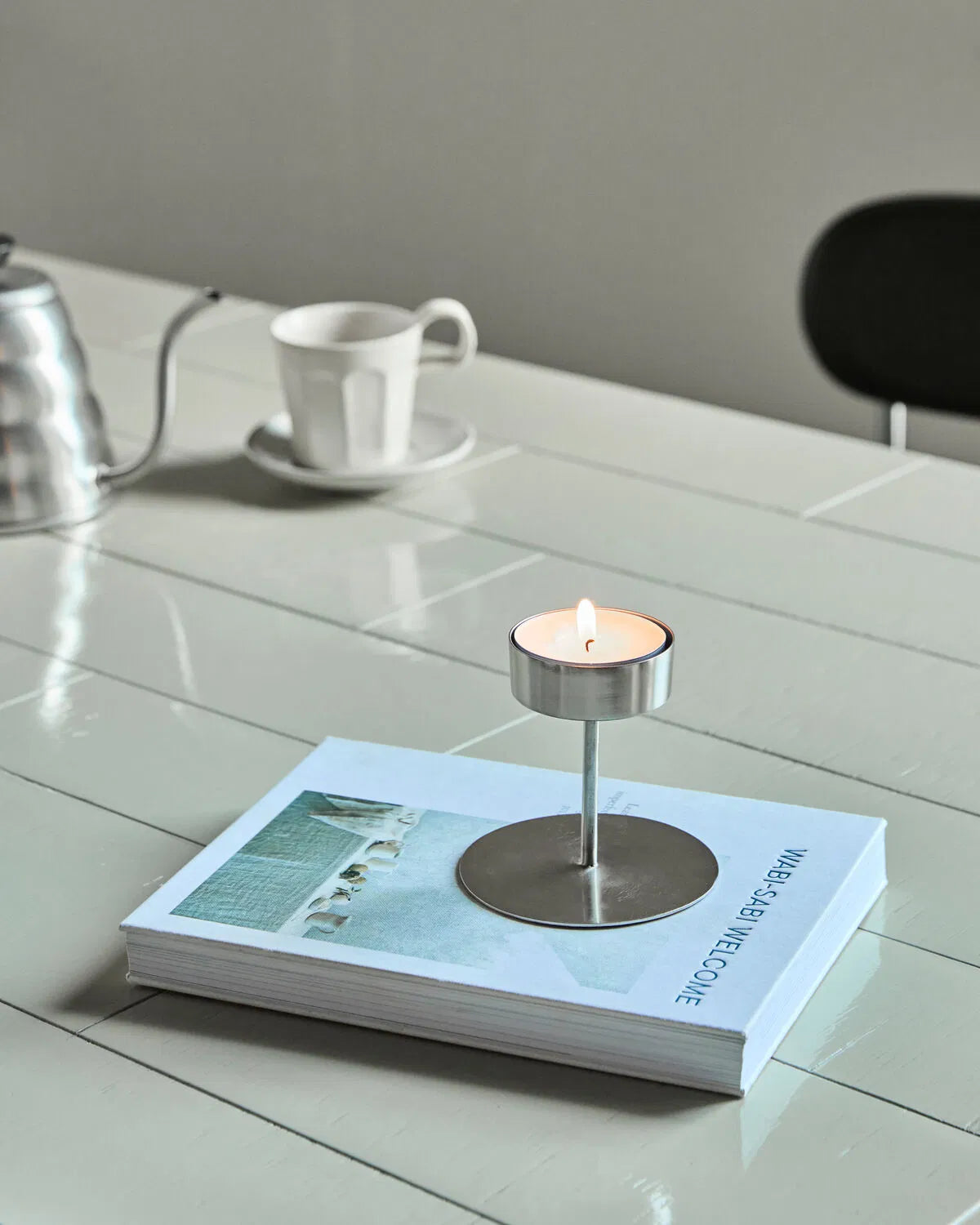 A silver Anit tealight holder is placed on a book, on a tiled table, with a lit tealight in it. In the background is a silver kettle and white mug.