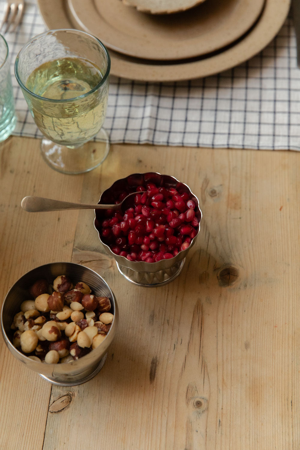 Stainless steel ice cups, fluted and plain, filled with pomegranate seeds and mixed nuts beside a goblet of white wine
