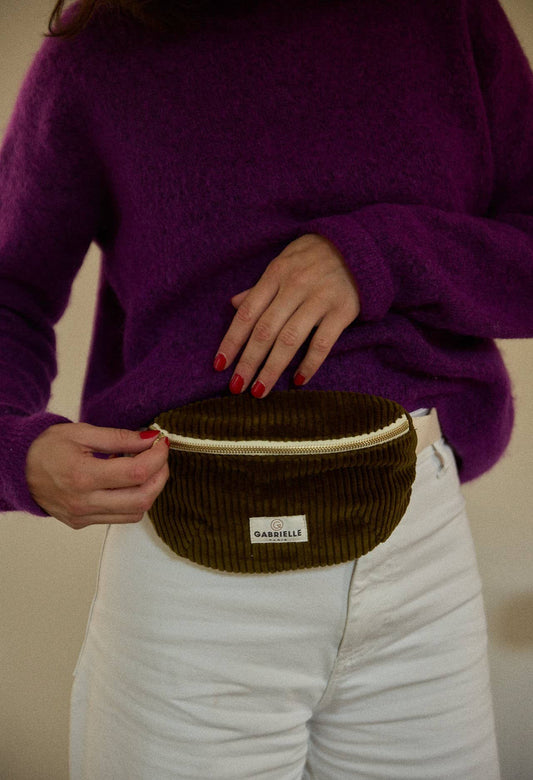 Person wearing a purple sweater and white pants with a small olive velvet corduroy waist bag around their waist.
