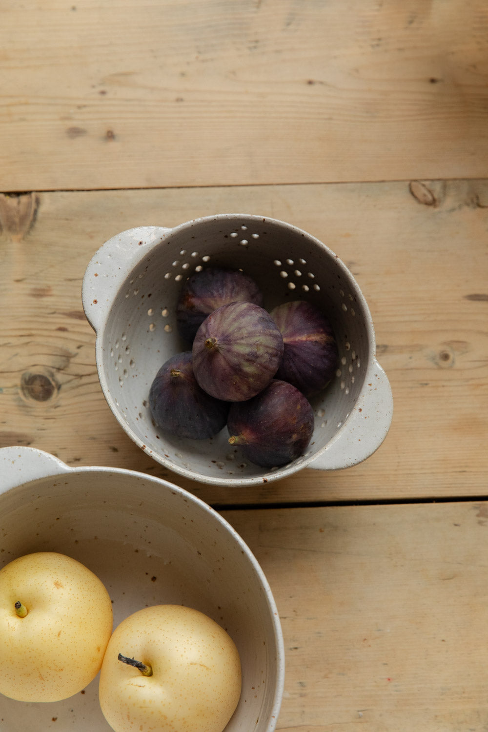 Speckled stoneware berry bowl of ripe figs beside a bowl of pears on a pine table