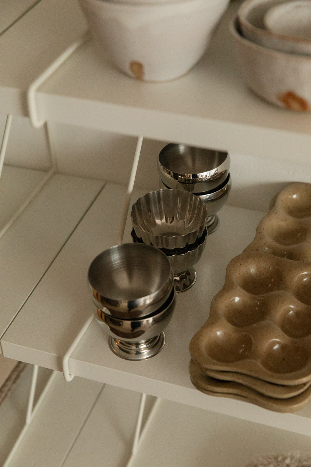 Stacked speckle stoneware egg trays and silver footed cups on a white shelf