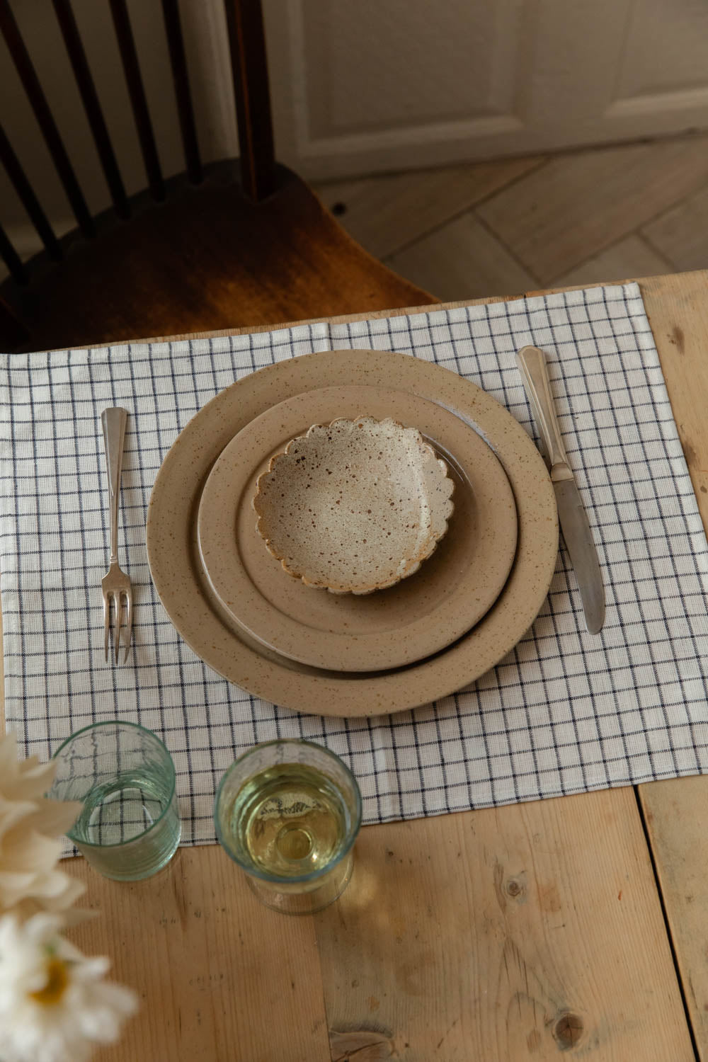 Overhead place setting with stacked speckled stoneware plates and a scalloped dish on a grid linen placemat, with recycled glass tumblers