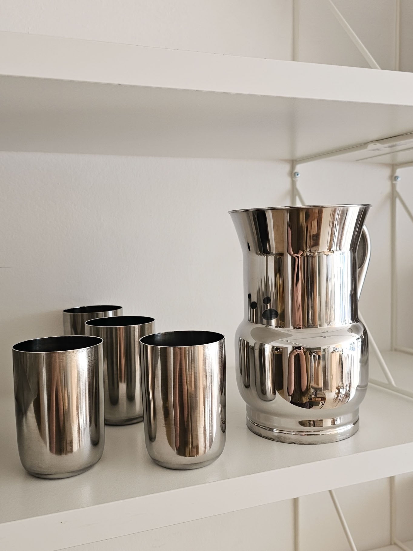 A polished stainless steel pitcher and four matching tumblers displayed on a white shelf.