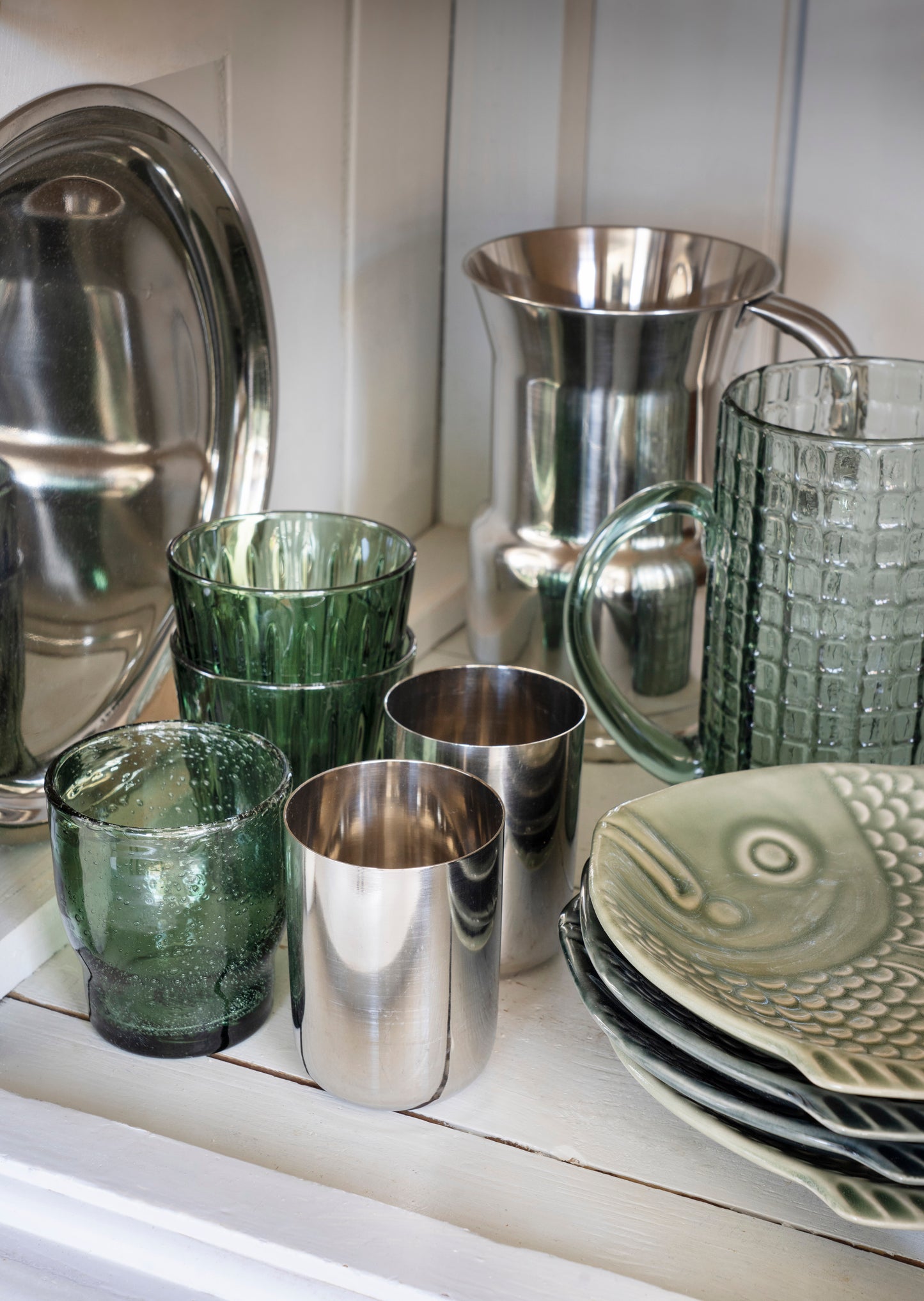 A collection of homeware and glassware displayed on a white shelf, including silver metal pitchers and tumblers, textured green glass cups, and a stack of gray ceramic plates with a fish design.