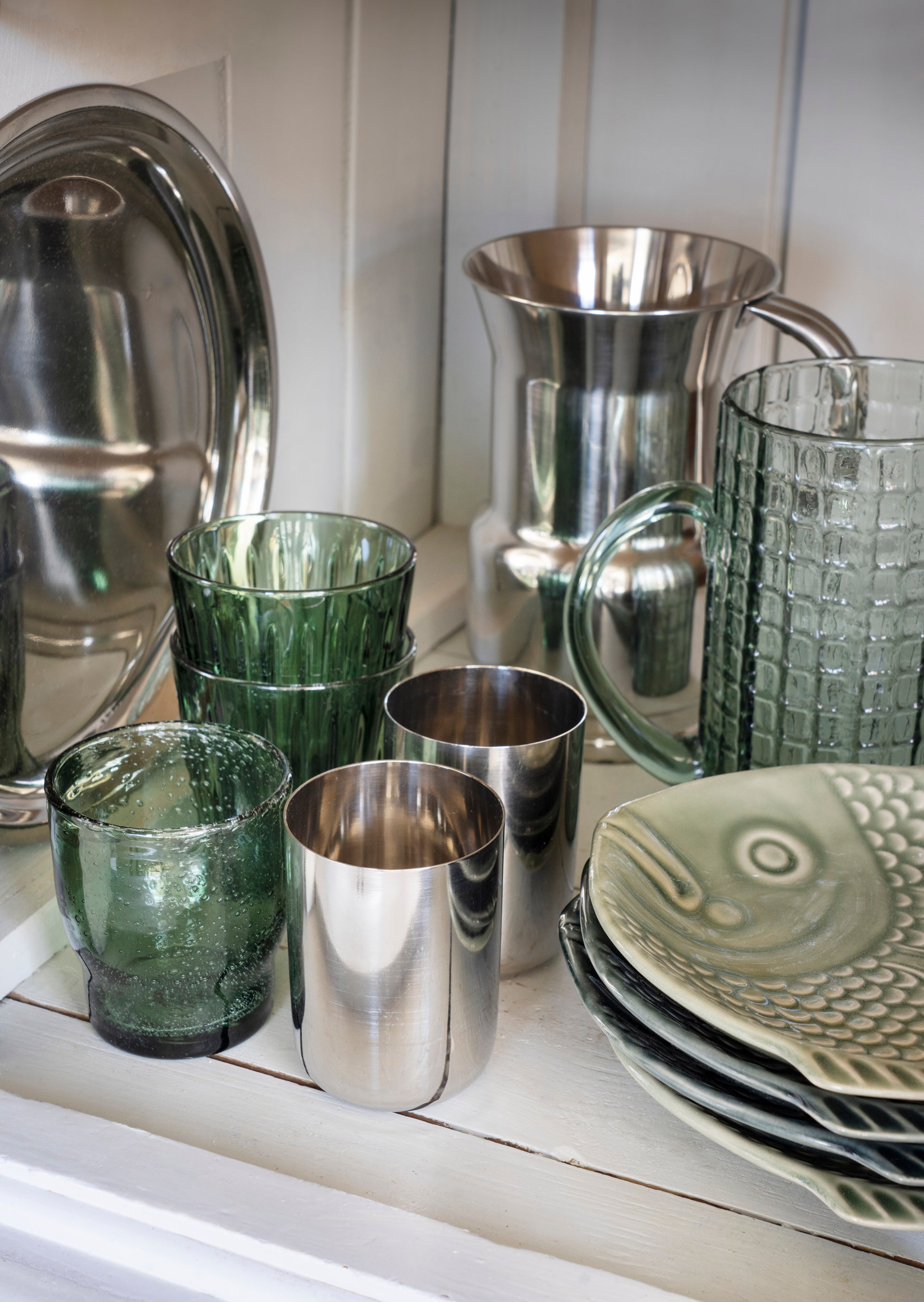 A collection of homeware and glassware displayed on a white shelf, including silver metal pitchers and tumblers, textured green glass cups, and a stack of gray ceramic plates with a fish design.