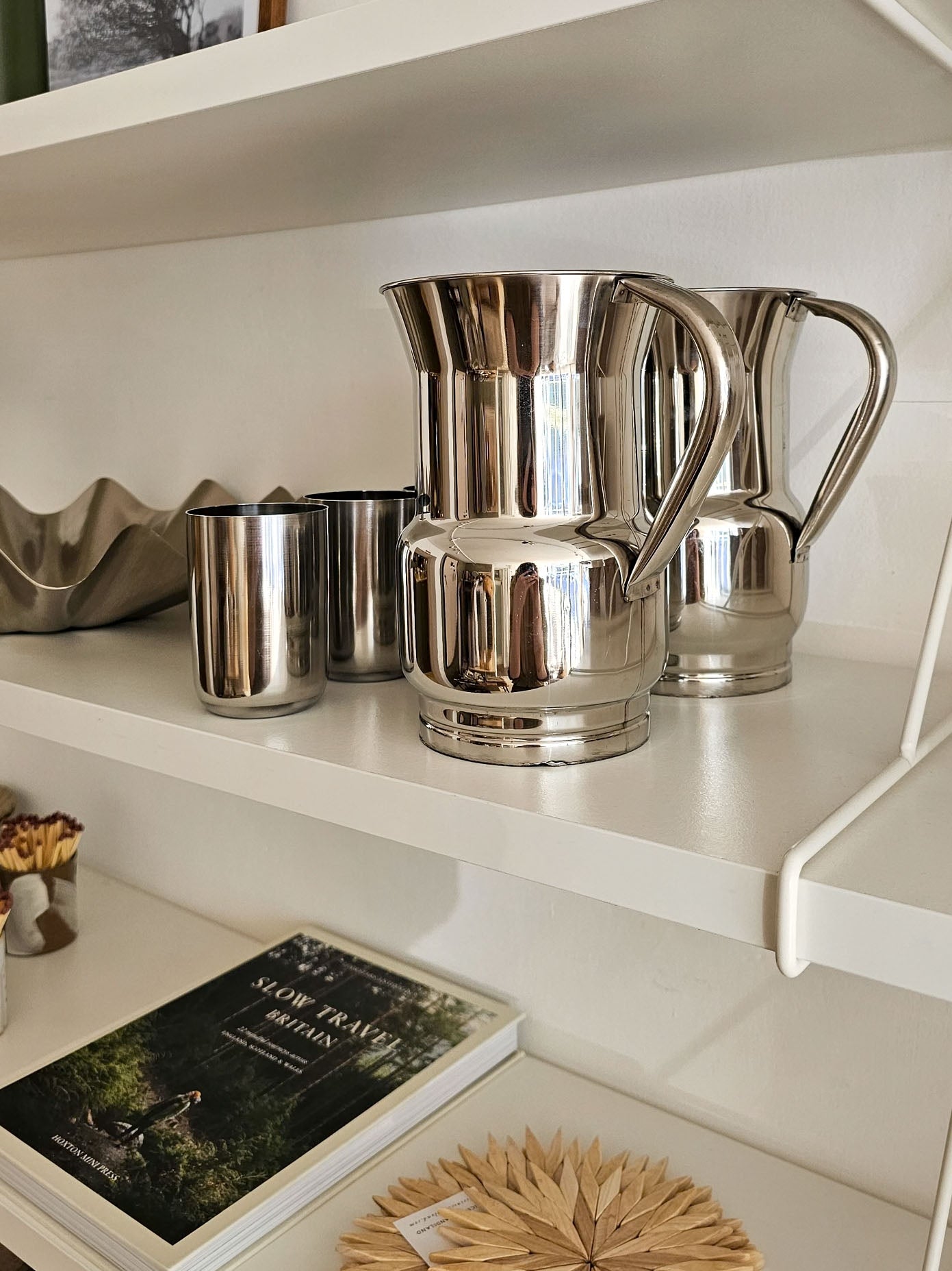 A polished stainless steel pitcher and matching metal cups displayed on white shelves in a home interior setting.