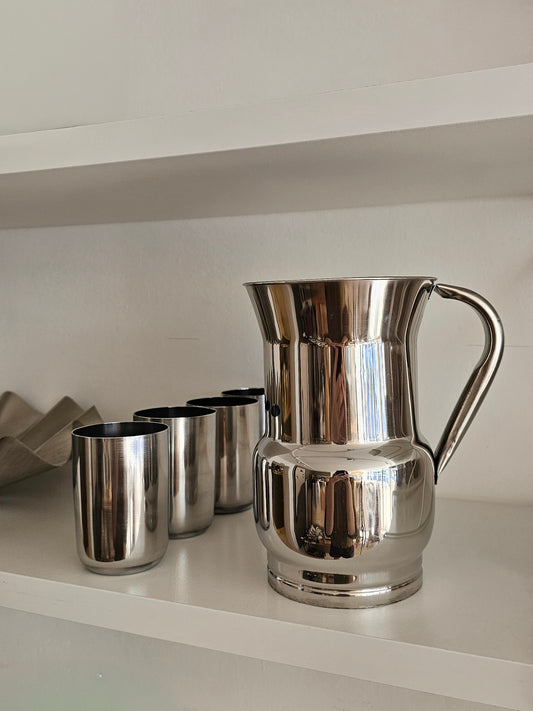 A polished stainless steel pitcher with a curved handle sits on a white shelf next to four matching metal tumblers against a white wall.