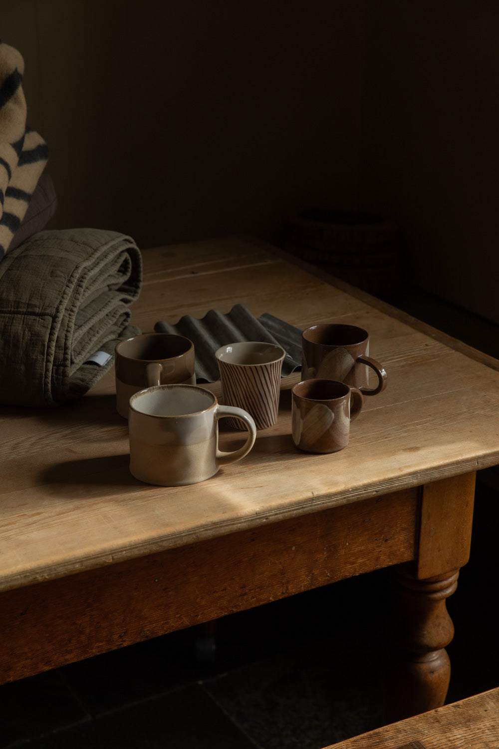 Half glazed and brushstroke stoneware mugs grouped on a wooden table beside a folded quilt