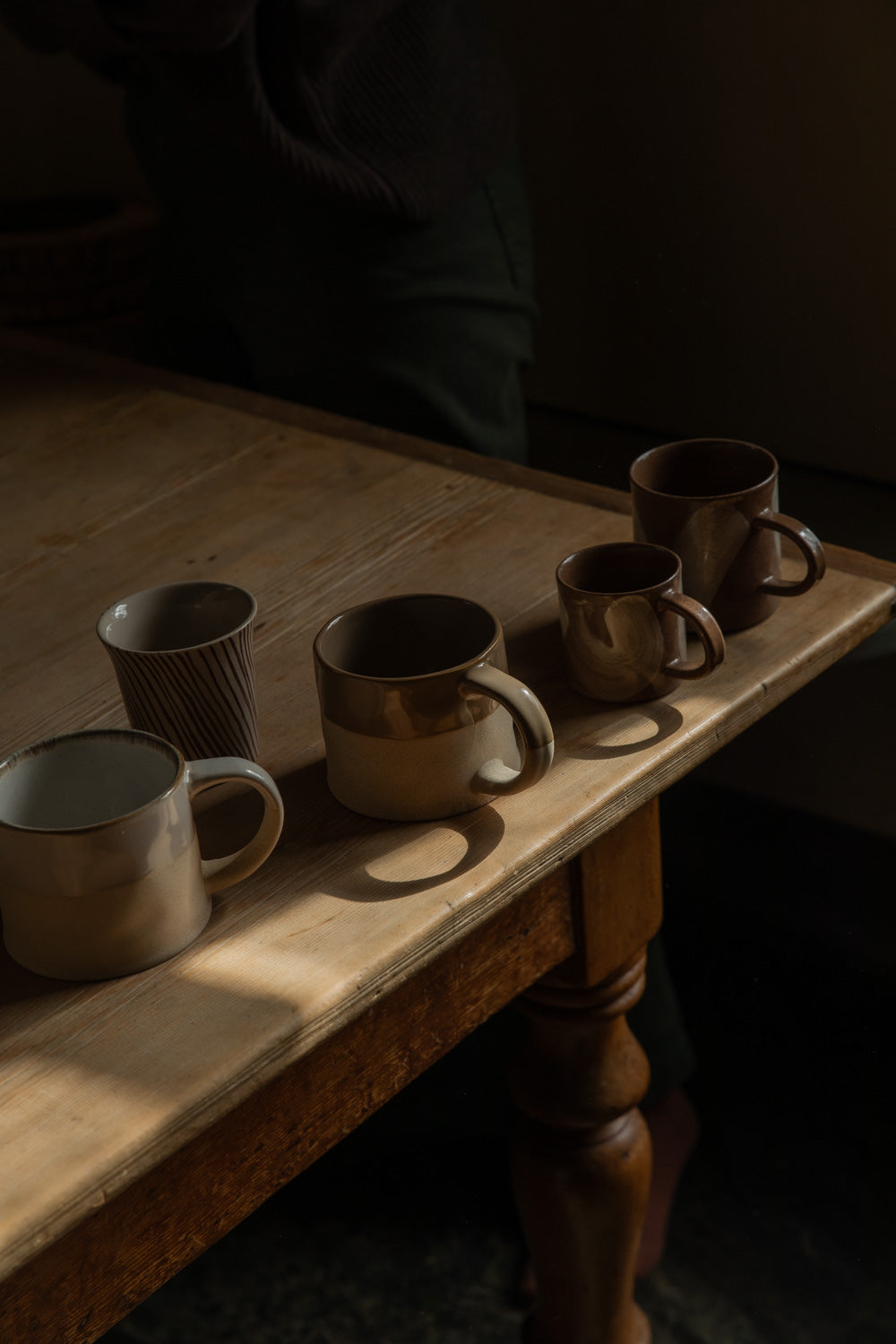 Handmade stoneware mugs in warm browns arranged on a scrubbed pine farmhouse table in low light