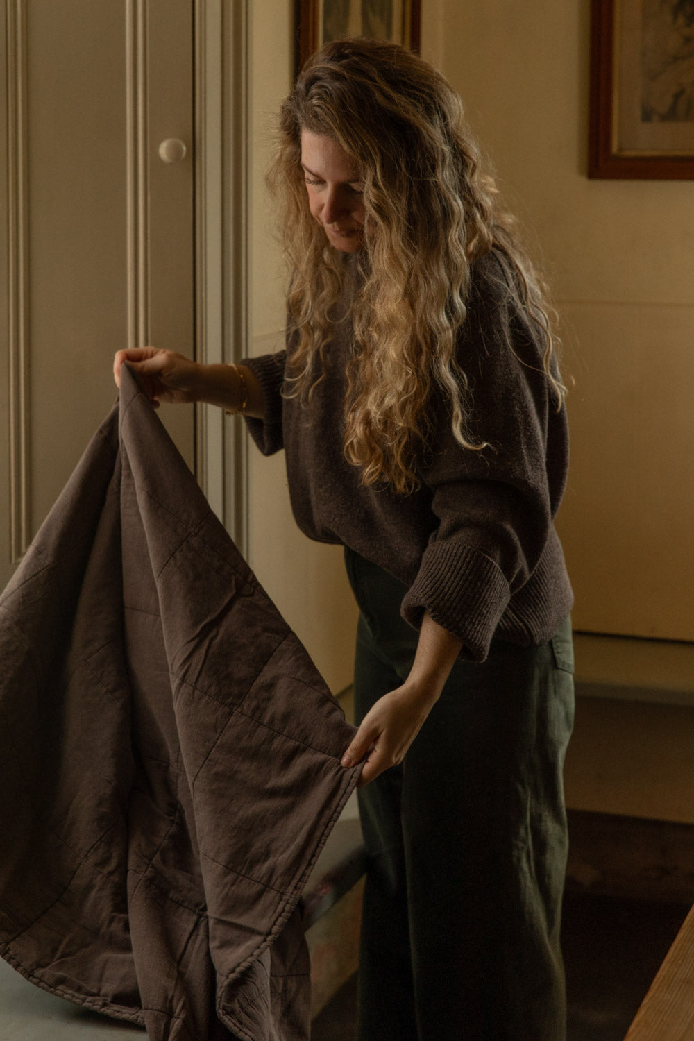 Woman shaking out a soft, earthy brown stonewashed cotton quilt in a panelled room