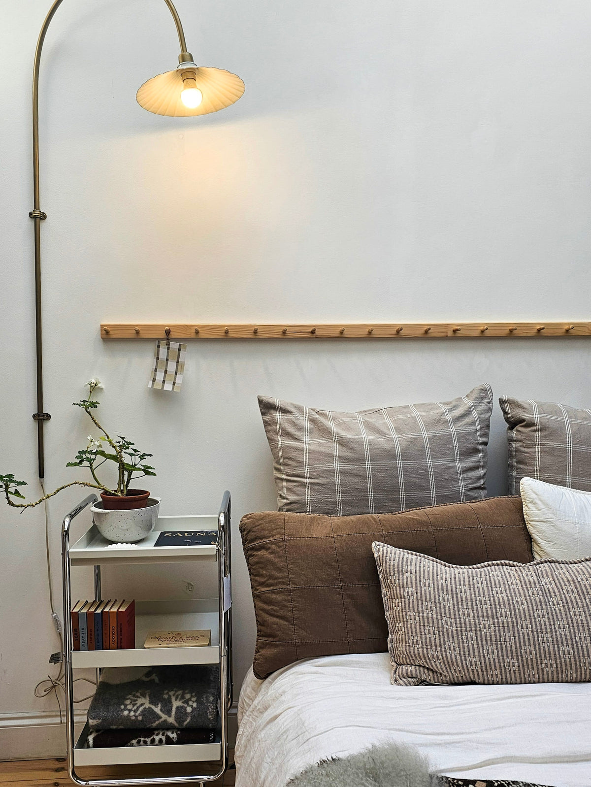 A cozy bedroom corner featuring a bed with brown and striped pillows, a wooden coat rack on the wall, a brass floor lamp, and a small metal side cart with books and a plant.