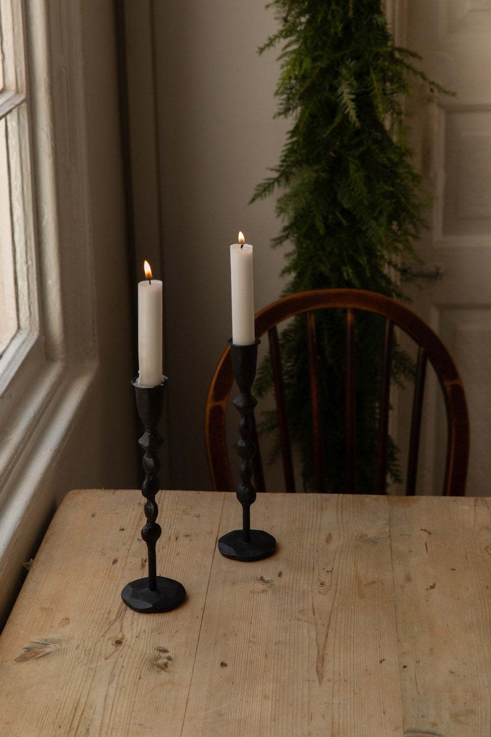 Two tall hand-forged black iron candle holders with lit white candles on a table by a window