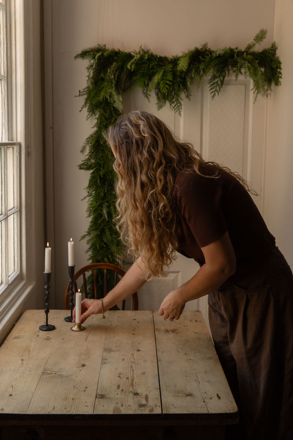 Woman arranging tall black iron and brass candle holders on a kitchen table beneath a foliage garland