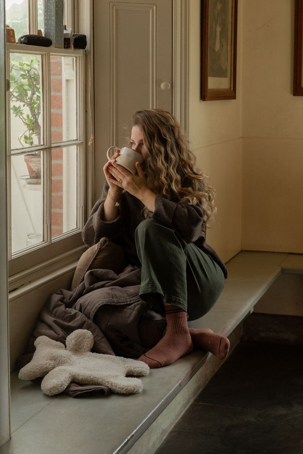 Woman sipping from a mug on a window seat, a teddy sheepskin hot water bottle and brown quilt beside her