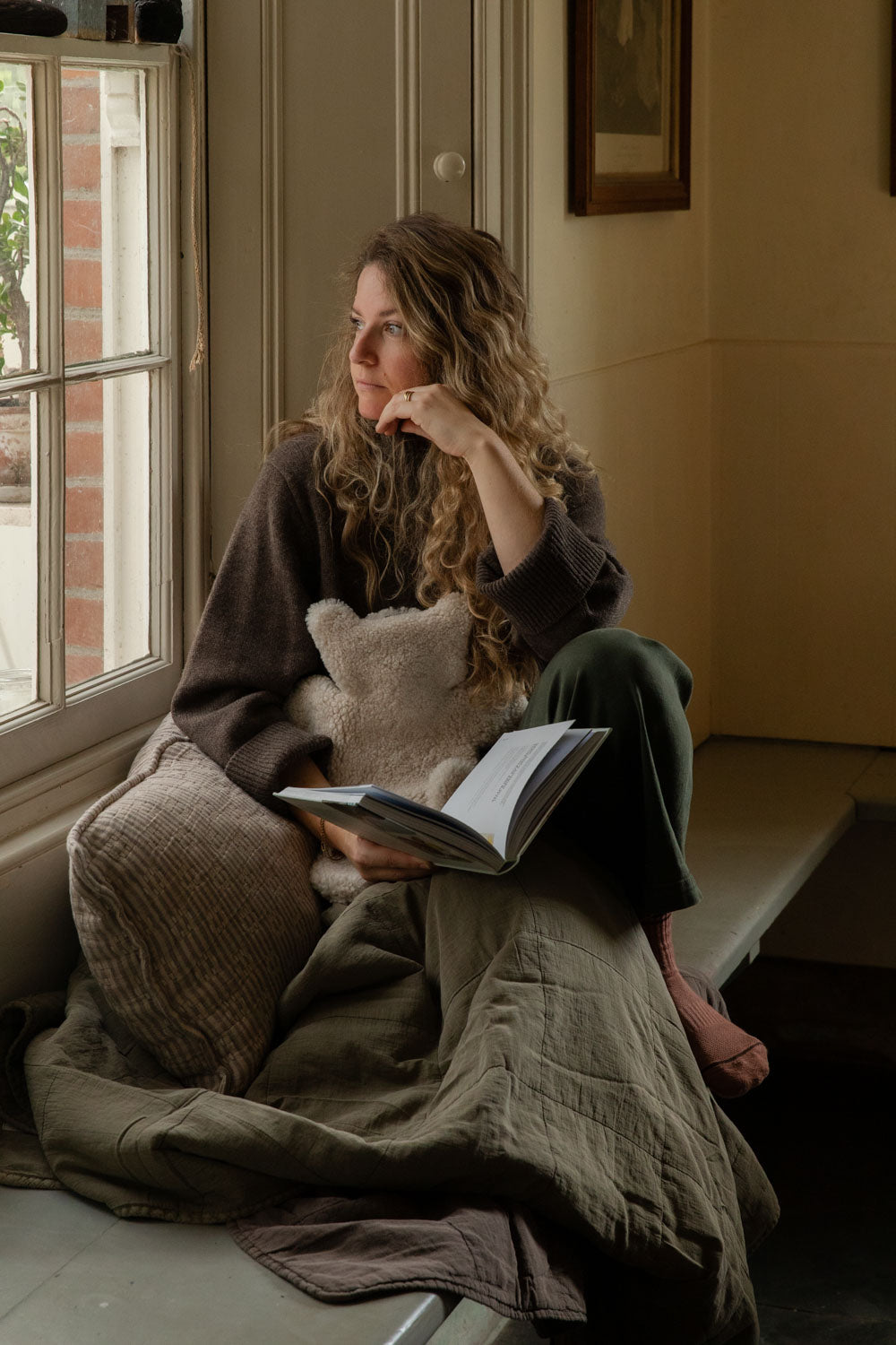 Woman reading on a window seat with a teddy sheepskin hot water bottle, a striped cushion and a green quilt