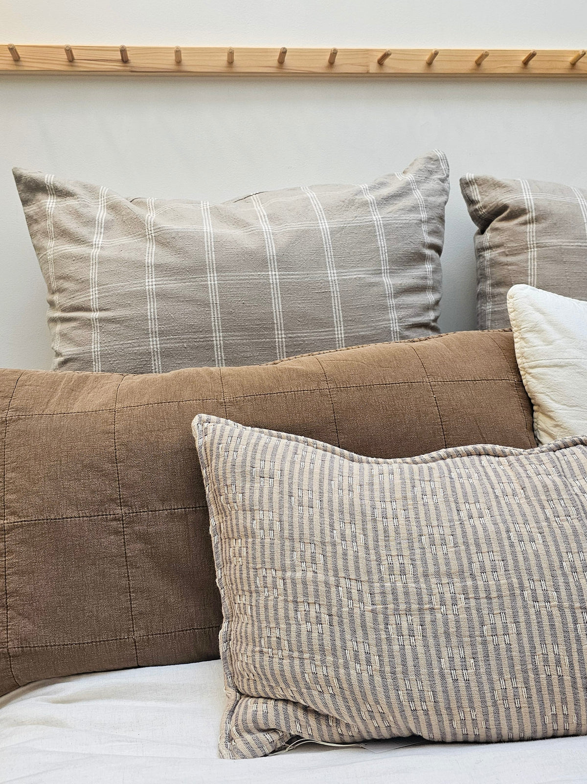 A styled collection of home decor pillows in neutral tones, including a large brown quilted pillow, a striped taupe pillow, a textured beige and gray striped lumbar pillow, and a white accent pillow, arranged against a light wall with a wooden headboard.