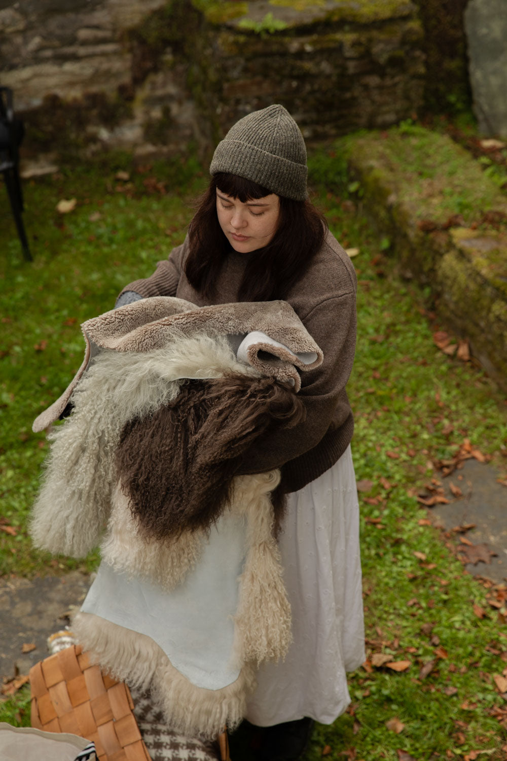 Woman in a green beanie carrying an armful of grey, brown and cream sheepskin rugs across a mossy garden