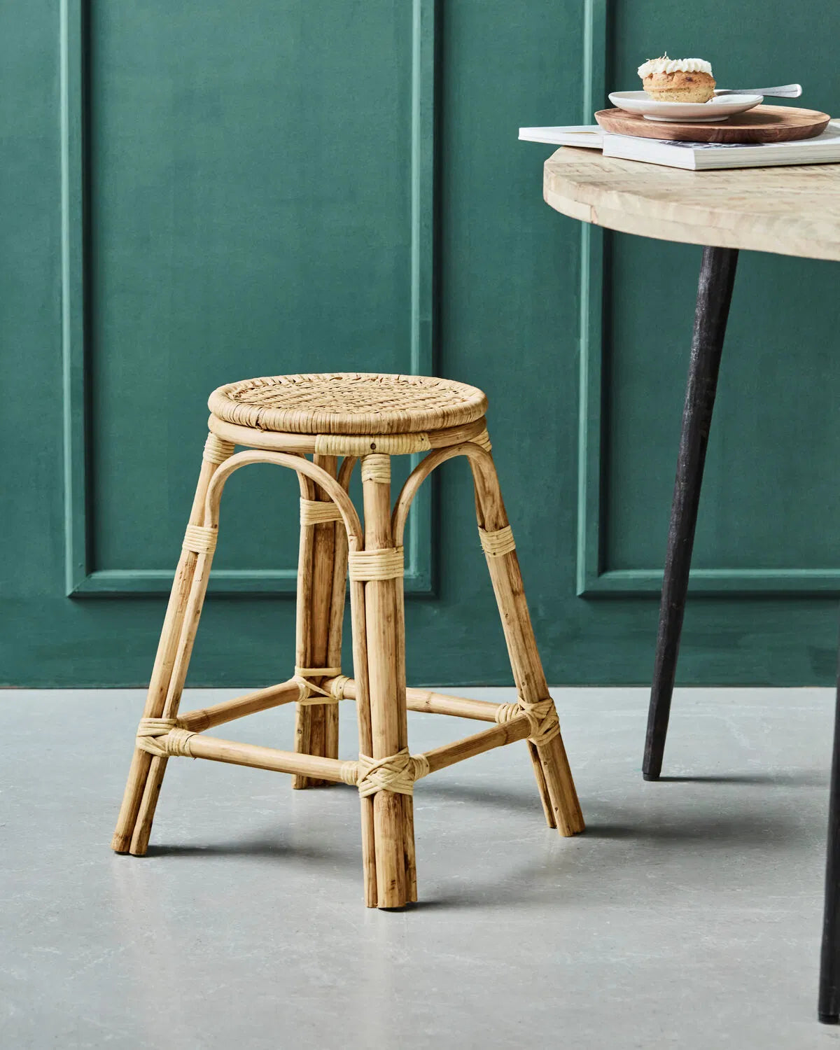 Trenza rattan stool shown against a blue/green wall, next to a table.