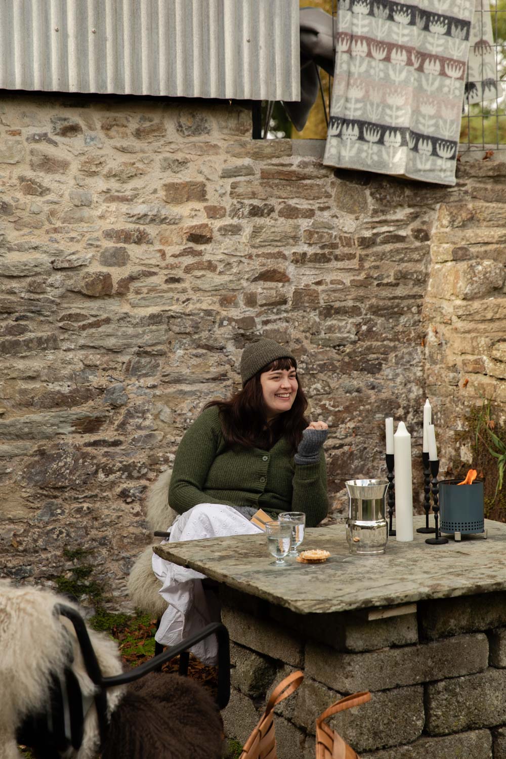 Friends at a stone garden table with candles and sheepskins, a grey and pink Tulip lambswool blanket hanging on the wall above