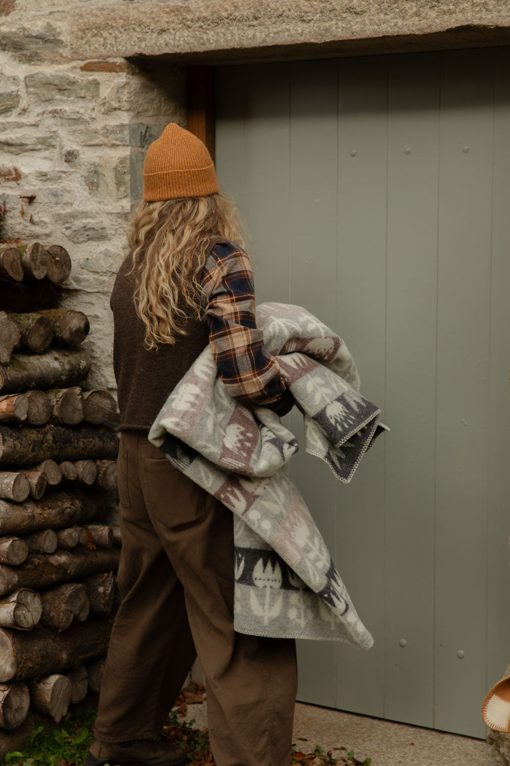 Woman in a check shirt and orange beanie carrying a folded grey and pink Tulip lambswool blanket past a log store