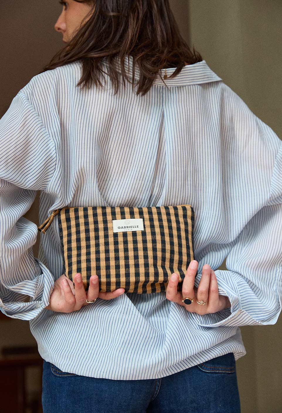 Person holding a checked Vichy Cafe cotton pouch behind their back against a neutral background.