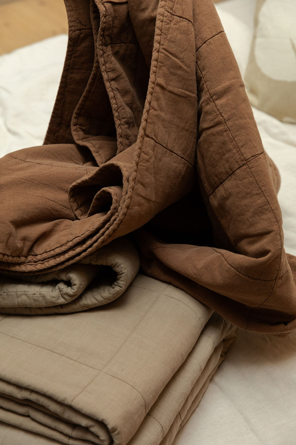Close-up of vintage camel cotton quilt draped over folded neutral colourway quilts on a white linen bed, showing blanket stitch edge and quilted block texture detail.