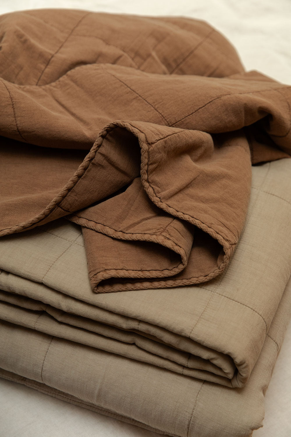 Vintage camel cotton quilt loosely draped over folded neutral colourway quilts on a white linen bed, showing soft quilted texture and blanket stitch edge detail up close.