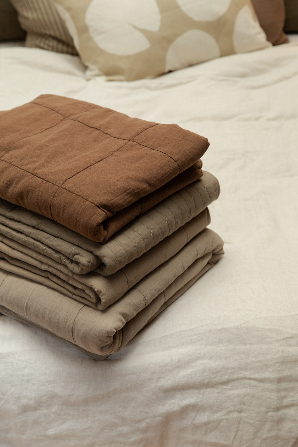 Cotton quilts in vintage camel, taupe and sand colourways stacked and folded on a white linen bed with Fine Little Day Water Lilies cushion in background.