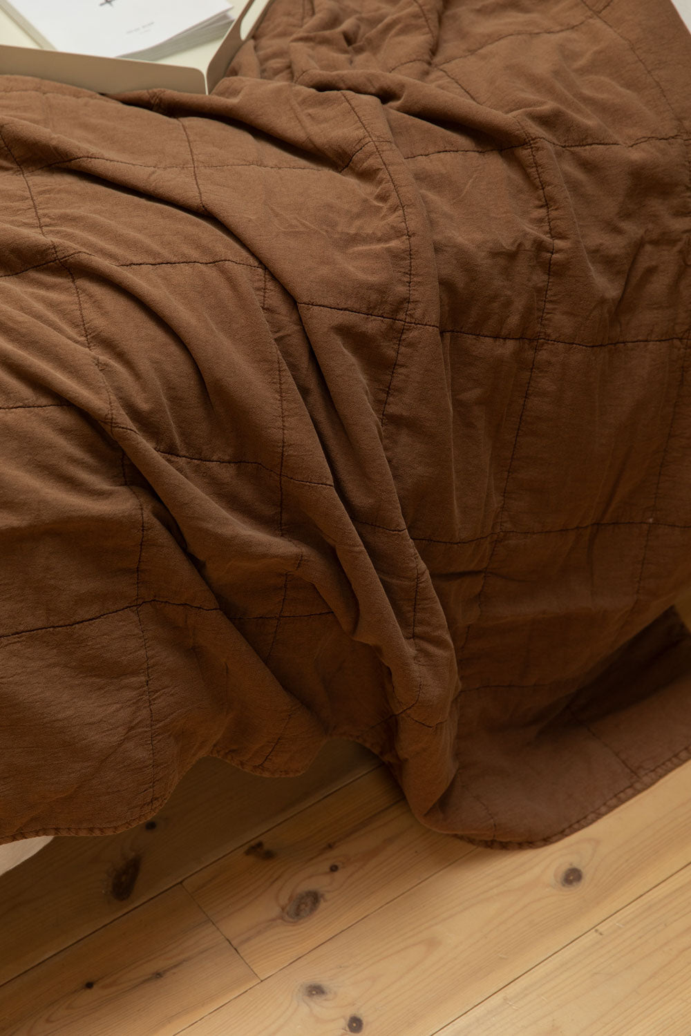 Close-up of vintage camel cotton quilt showing quilted block texture detail draped over the end of a bed with pine floorboards below.