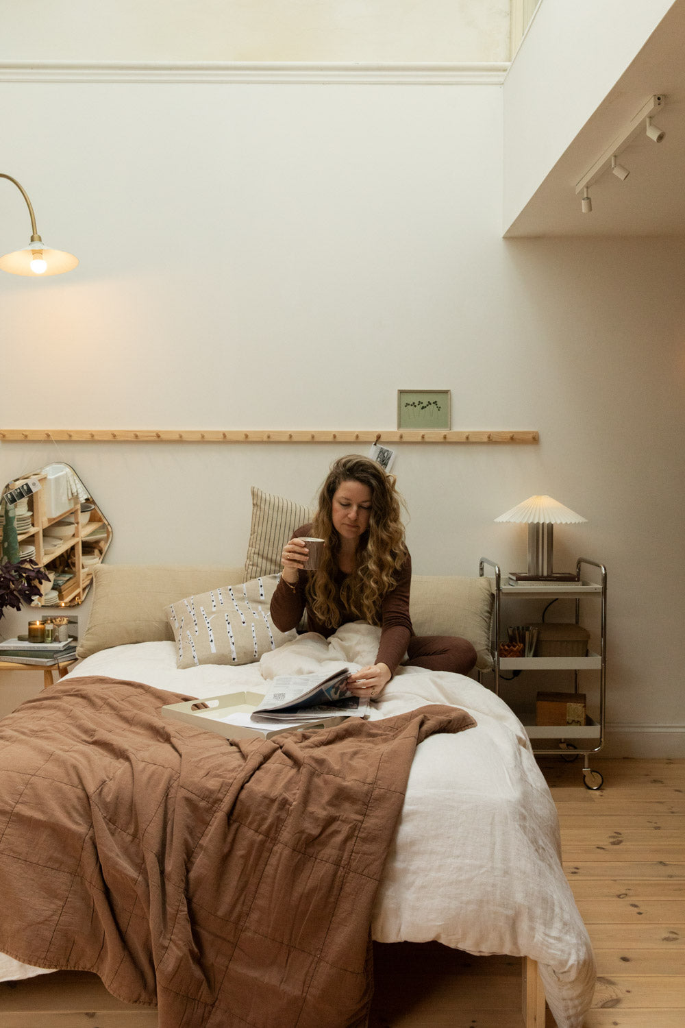 Woman sitting in bed with coffee and magazine surrounded by vintage camel cotton quilt, Blomus Taite pleated table lamp and pine peg rail.