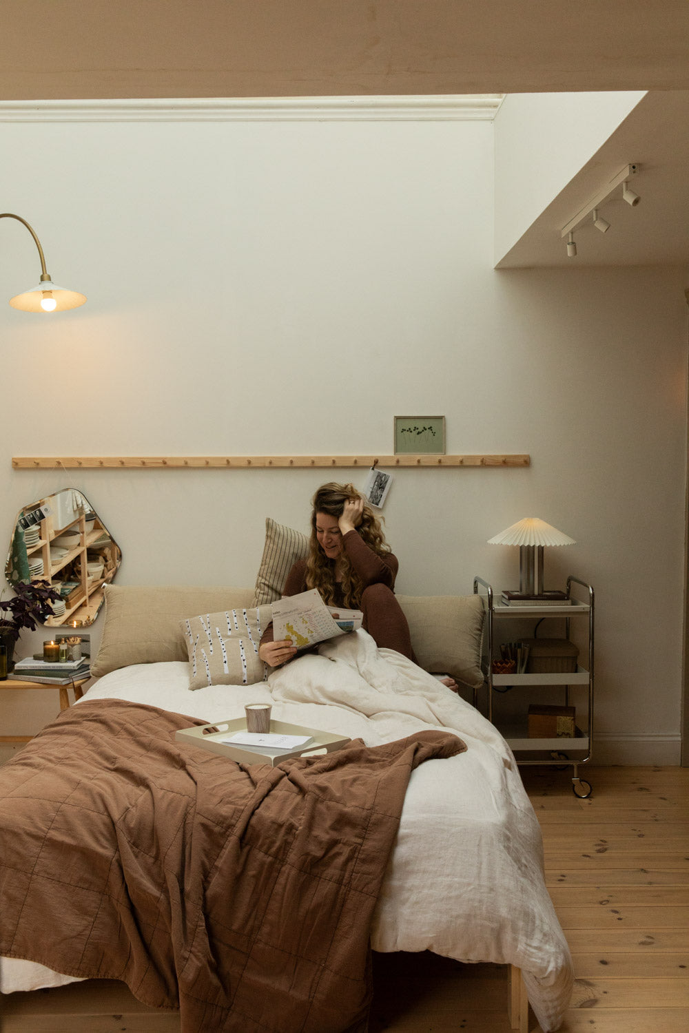 Woman sitting up in bed reading with vintage camel cotton quilt, arc brass floor lamp, Blomus Taite pleated table lamp and skylight above.