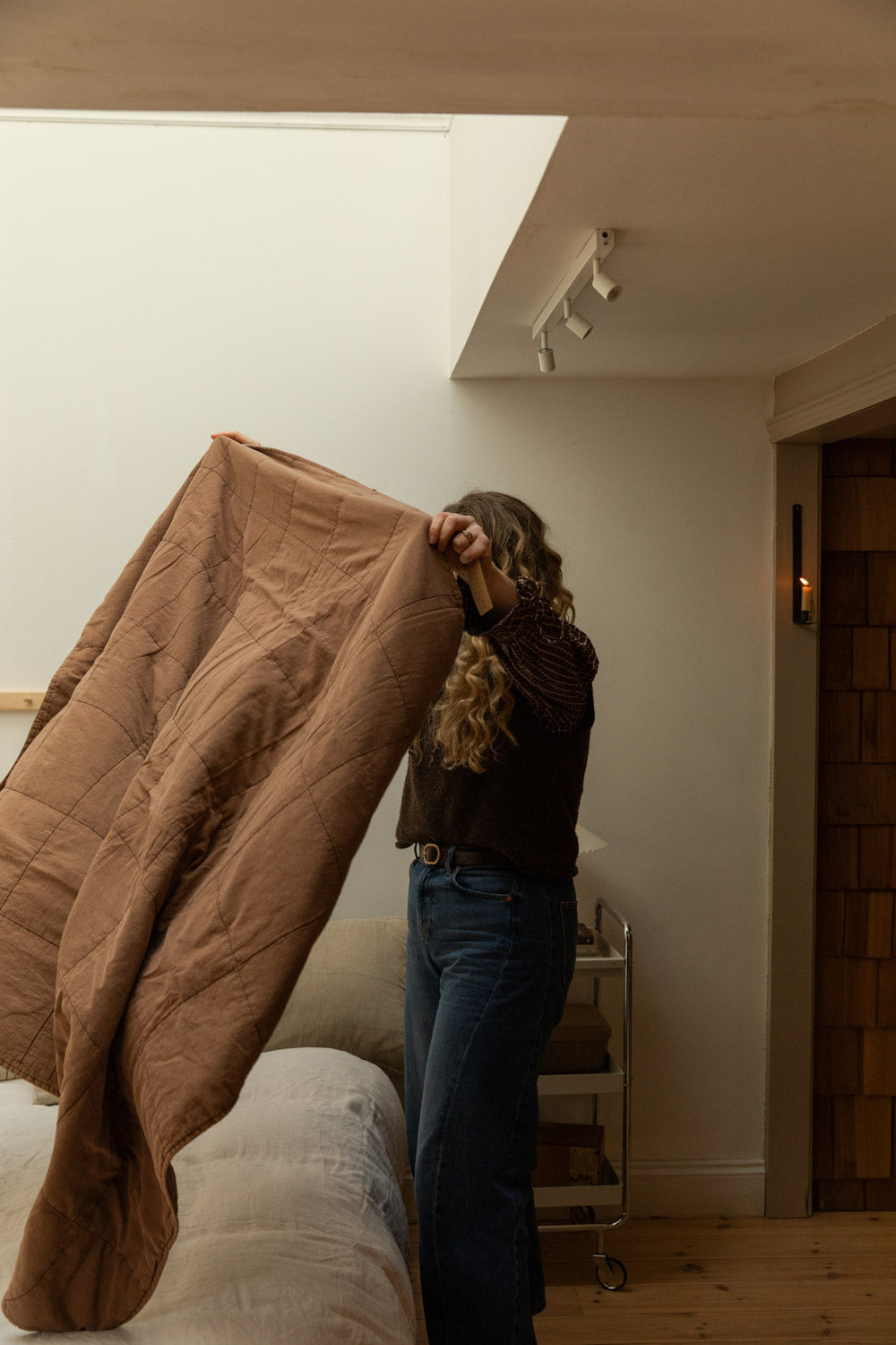 Woman shaking out vintage camel cotton quilt beside a bed in a high-ceilinged bedroom with skylight and chrome trolley bedside table.