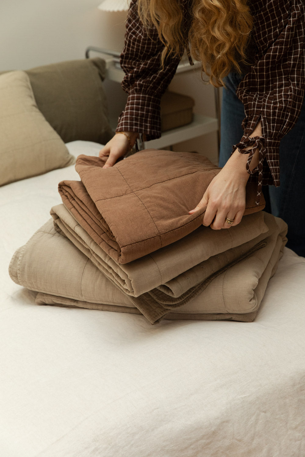 Woman stacking folded cotton quilts in multiple colourways including vintage camel on a white linen bed.
