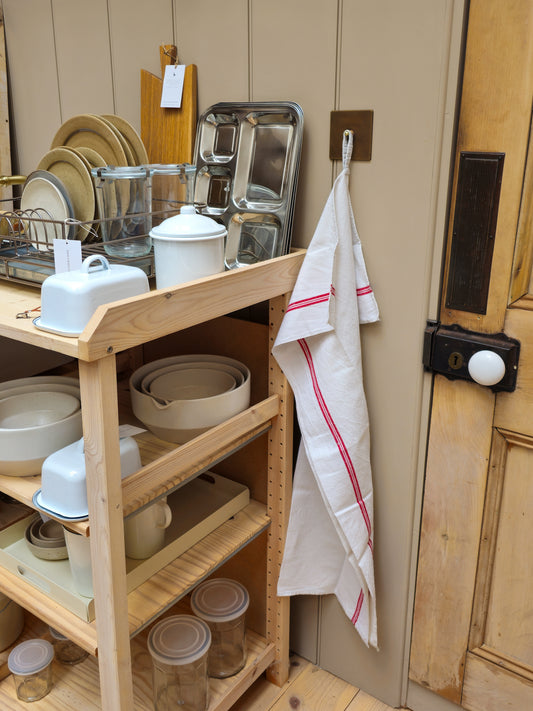 A light natural wood kitchen shelving unit holding white ceramic dishes, glass jars, and metal cookware, with a white kitchen towel hanging from a hook on the beige paneled wall beside a wooden door.