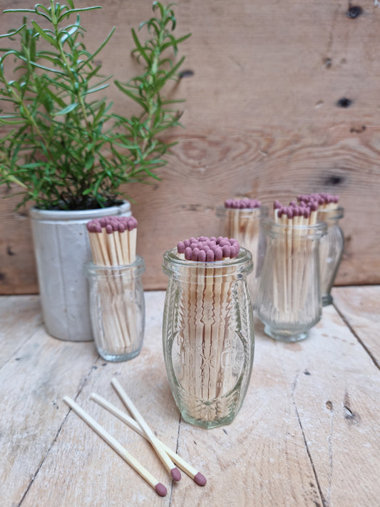 Vintage glass match pots with long matches placed on a wooden surface, with a potted plant in the background.